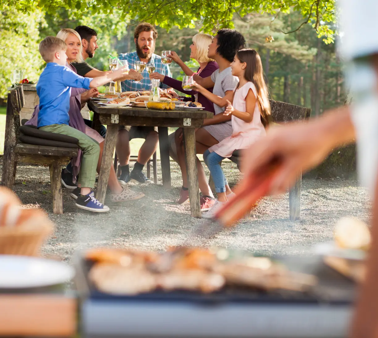 Gesellige Runde mit Freunden und Familie beim Essen an einem Gartentisch, im Vordergrund unscharf ein Grill – passendes Ambiente für Sommerzubehör für Männer und BBQ-Werbegeschenke.